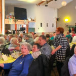 Eine Feier mit vielen Seniorinnen und Senioren, sie sitzen an langen Tischen und lauschen dem musikalischen Programm.