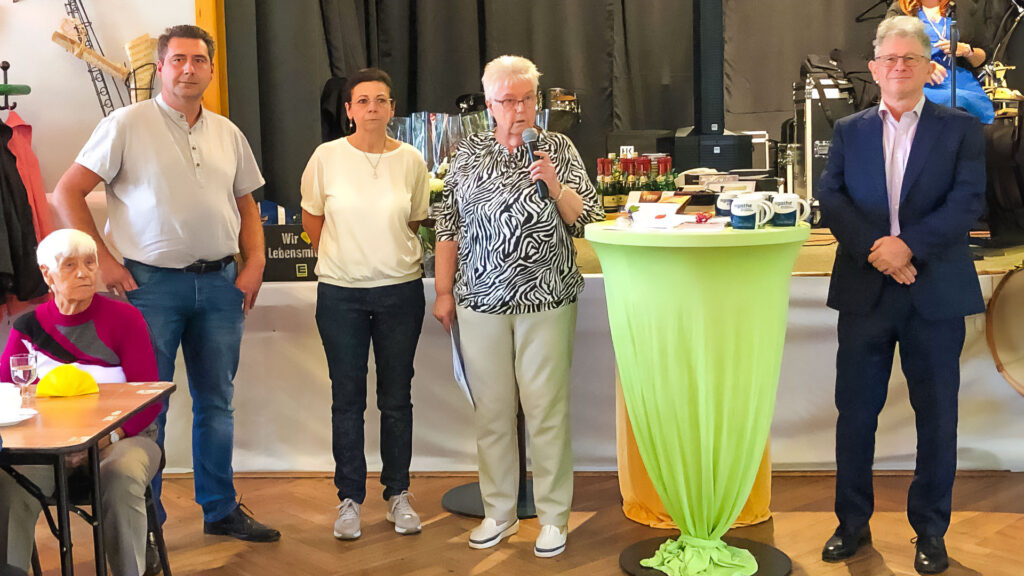 Zwei Männer und zwei Frauen, eine davon mit grauen Haaren und einem Mikrofon, stehen an einem grünen Tisch, direkt vor einer Bühne.