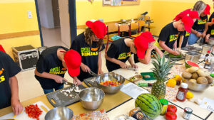 Kinder einer fünften Klasse, gekleidet mit schweren T-Shierts, stehen an einem großen Tisch und bereiten Zutaten für das gemeinsame Grillen vor.