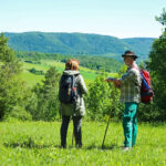 Zwei wandernde Personen auf einer grünen Wiese, im Hintergrund sind waldbedeckte Berge zu sehen.