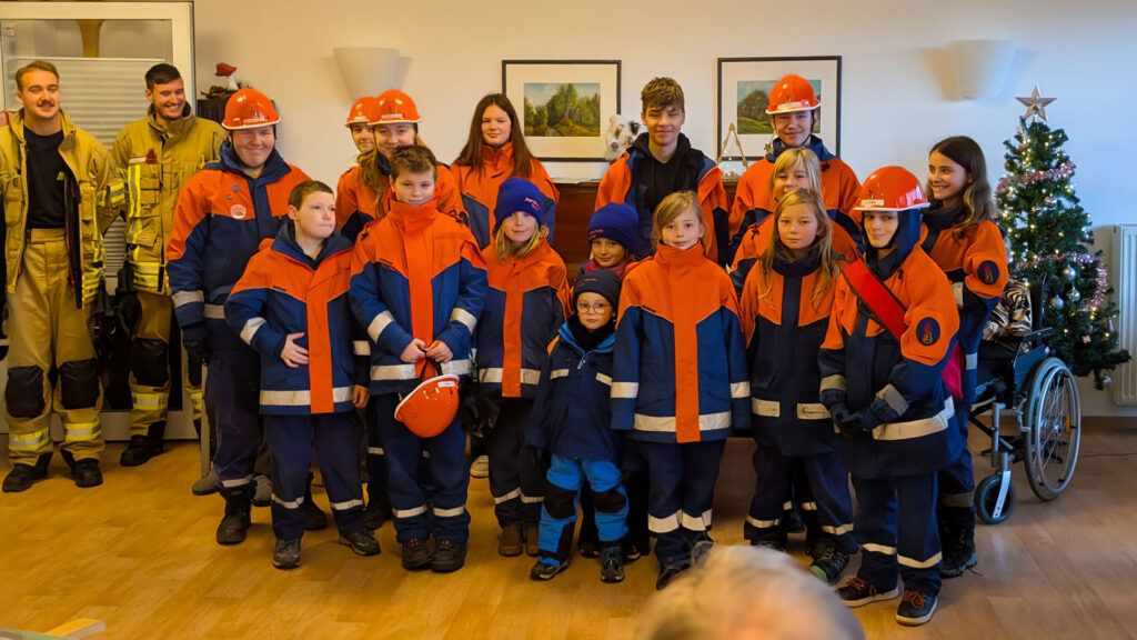 Kinder in Feuerwehrkleidung und zwei erwachsene Feuerwehrleute stehen neben einem Weihnachtsbaum für ein Foto im Altersheim bereit