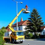 Das Bild zeigt einen gelben Kranwagen mit einem Arbeiter auf einer Kranplattform, der an einer Straßenlaterne arbeitet. Die Szene spielt vor einem klaren blauen Himmel, mit Wohnhäusern und Bäumen im Hintergrund. (KI)