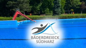 Das Bild zeigt ein lebhaftes Freibad mit einer roten Rutsche und dem Logo von „Bäderdreieck Südharz“ darüber. Das Schwimmbad ist von Grünflächen und parkähnlichen Einrichtungen umgeben.