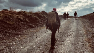 Das Bild zeigt eine Gruppe von Wanderern, die auf einem zerklüfteten Schotterweg in einer bergigen Gegend wandern. Der Himmel ist mit Wolken bedeckt, was auf einen kühlen und möglicherweise windigen Tag hindeutet.