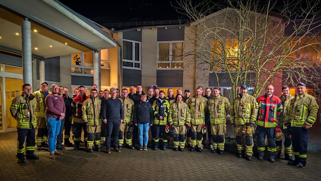 Das Bild zeigt eine große Gruppe von Feuerwehrleuten in Uniform, die vor einem Gebäude stehen und für ein Gruppenfoto posieren. Im Hintergrund ist ein mehrstöckiges Gebäude bei Nacht zu sehen. Einige Zivilisten stehen ebenfalls in der Gruppe.