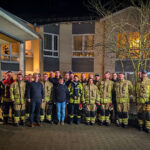 Das Bild zeigt eine große Gruppe von Feuerwehrleuten in Uniform, die vor einem Gebäude stehen und für ein Gruppenfoto posieren. Im Hintergrund ist ein mehrstöckiges Gebäude bei Nacht zu sehen. Einige Zivilisten stehen ebenfalls in der Gruppe.