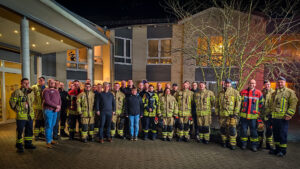 Das Bild zeigt eine große Gruppe von Feuerwehrleuten in Uniform, die vor einem Gebäude stehen und für ein Gruppenfoto posieren. Im Hintergrund ist ein mehrstöckiges Gebäude bei Nacht zu sehen. Einige Zivilisten stehen ebenfalls in der Gruppe.