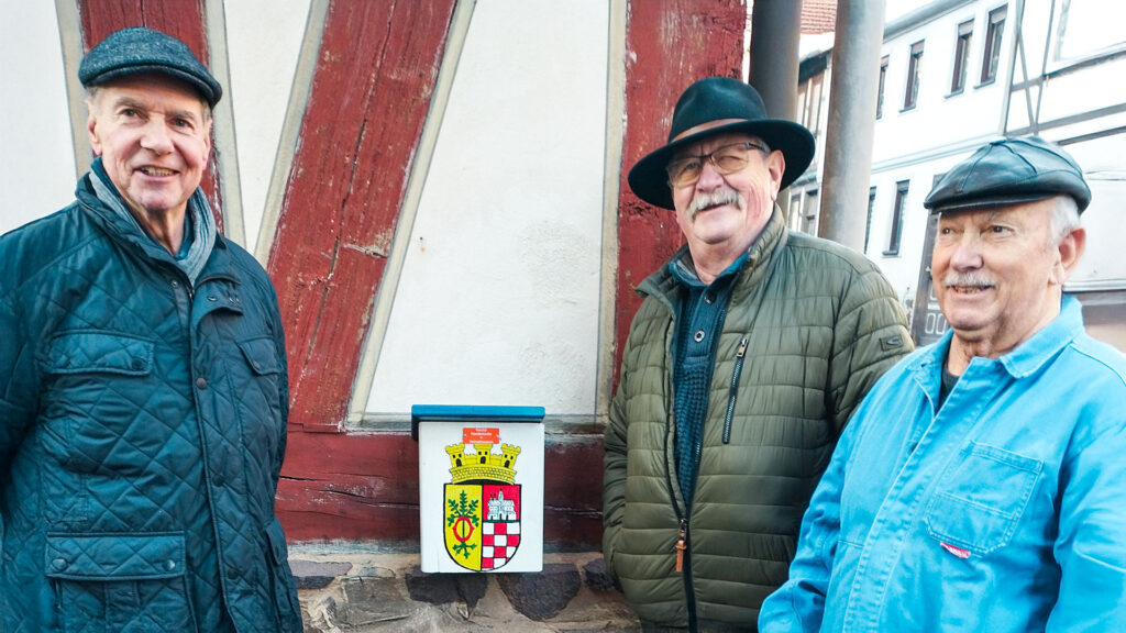 Das Foto zeigt drei ältere Herren vor einem Gebäude. Sie stehen vor einem kleinen Schild, das ein Wappen zeigt. Die Männer tragen verschiedene Mützen und Jacken. Das Gebäude scheint aus Holz zu sein und hat eine rote Farbe. Der Hintergrund deutet auf eine historische Stadtlandschaft hin.