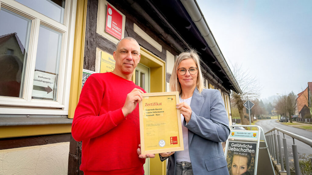 Das ist ein Foto von zwei Personen vor einem Gebäude, die ein Zertifikat halten. Ein Mann und eine Frau stehen vor einem Fachwerkhaus und halten gemeinsam ein großes Zertifikat, vermutlich eine Auszeichnung oder eine Urkunde, hoch.