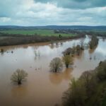 Das Bild zeigt eine Überschwemmung in einer ländlichen Gegend, wahrscheinlich aus der Vogelperspektive aufgenommen, möglicherweise mit einer Drohne. Ein Fluss oder ein Bach ist über seine Ufer getreten und hat weite Teile des umliegenden Ackerlandes überflutet. Das Wasser erscheint schlammig und braun, was auf eine beträchtliche Trübung hindeutet, möglicherweise durch den aufgewirbelten Boden. An einigen Stellen sind überflutete Bäume und Vegetation zu sehen.