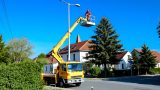 Das Bild zeigt einen gelben Kranwagen mit einem Arbeiter auf einer Kranplattform, der an einer Straßenlaterne arbeitet. Die Szene spielt vor einem klaren blauen Himmel, mit Wohnhäusern und Bäumen im Hintergrund. (KI)